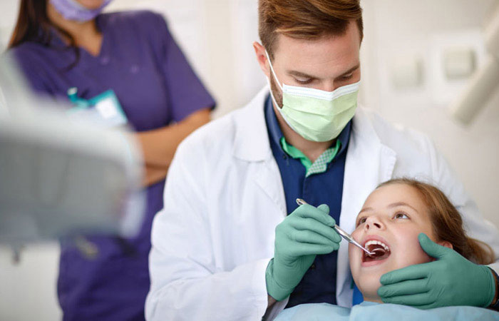 A dental hygienist with protective gloves is performing a dental procedure on a child while being assisted by another person, all wearing medical attire.