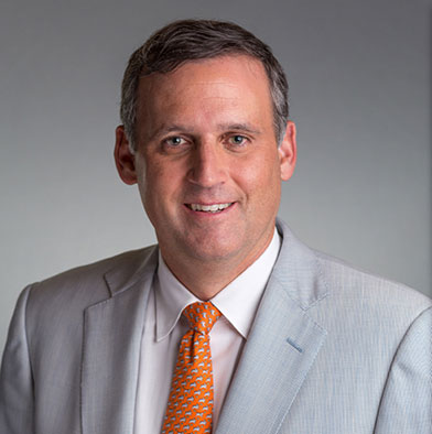 The image shows two photographs side by side of a man wearing a suit with a tie, smiling at the camera. He has short hair, and the background is neutral, focusing attention on him.