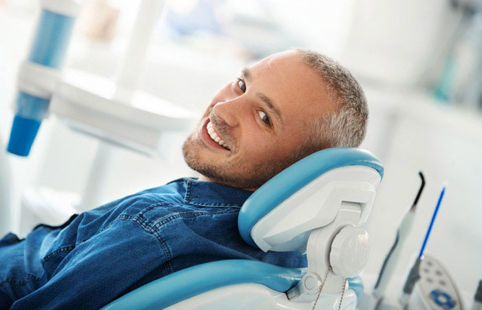 The image shows a man sitting in a dental chair with his eyes closed, wearing a blue shirt and jeans, smiling slightly, and looking directly at the camera.