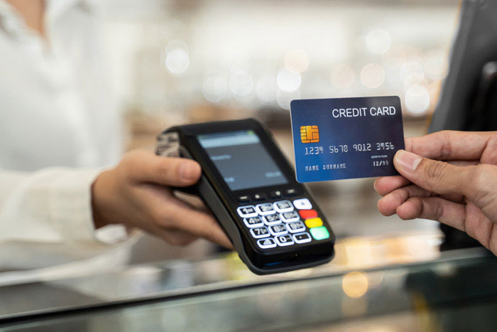 The image shows two hands holding a credit card near a card reader at a cash register, with a person standing behind the counter.