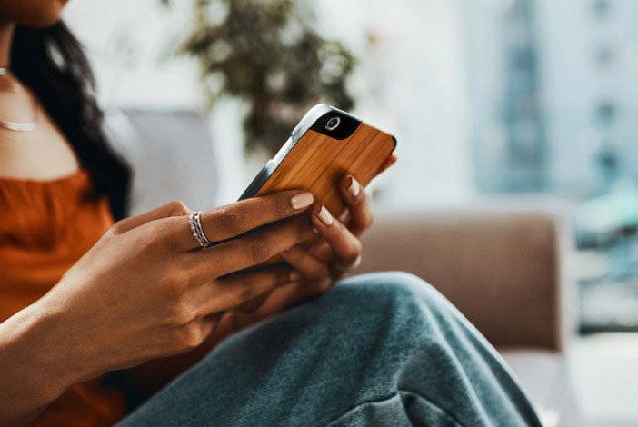 This is a photograph showing a person holding a smartphone with both hands while seated on a couch, using the device with focus. The individual appears relaxed and engaged with their phone s content.